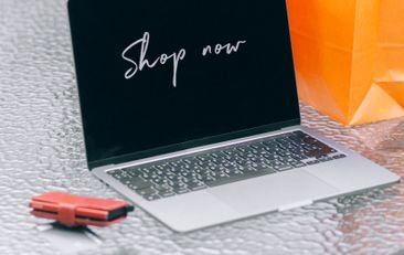 Laptop displaying 'Shop now' with vibrant shopping bags on glass table outdoors.